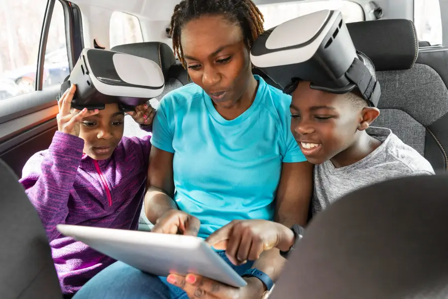 A woman and two children sit in the back seat of a car, engaged with a tablet device together.