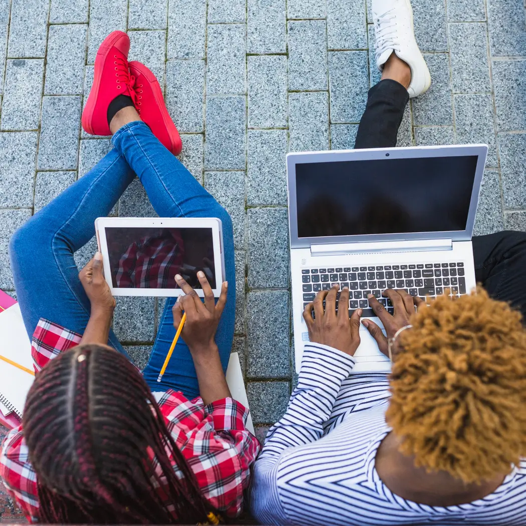 Two individuals seated on the ground, each using a laptop, engaged in a collaborative work session outdoors.